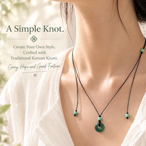 May include: A necklace featuring a dark green circular stone pendant suspended on a thin black cord. The cord includes three small turquoise-colored knotted accents and text that reads: A Simple Knot. Create Your Own Style. Crafted with Traditional Korean Knots. Carry Hope and Good Fortune.