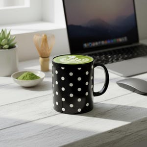 Pode incluir: Uma caneca preta com bolinhas brancas cheia de matcha latte, colocada sobre uma superfície de madeira branca. Um laptop, pó de matcha e um batedor também são visíveis. O conteúdo da caneca tem um padrão decorativo.