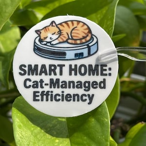 May include: A circular white sticker featuring a graphic of an orange tabby cat sleeping on a light blue robotic vacuum cleaner. Below the illustration, the text reads: SMART HOME: Cat-Managed Efficiency in bold, black, sans-serif capital letters.