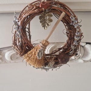 May include: A circular wreath made of brown twisted twigs, decorated with dried lavender sprigs, a small straw broom with a wooden handle, a metallic leaf charm, a reddish-brown stone, and a dark round seed pod, all suspended by a thin twine loop.