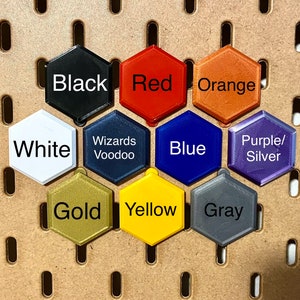 May include: A set of 10 hexagonal tiles in various colors, including black, red, orange, white, blue, purple, gold, yellow, and gray. Each tile has a different color and a word printed on it.