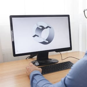 Może przedstawiać: Monitor komputera wyświetla model 3D szarej, okrągłej części mechanicznej. Osoba używa klawiatury i myszy na drewnianym biurku. Monitor jest czarny, a tło jasne.