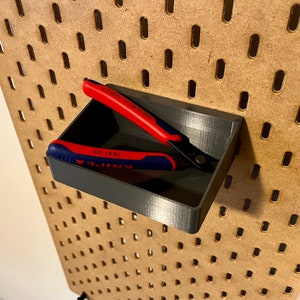 May include: A gray plastic tool holder with two red and blue pliers inside. The holder is mounted on a pegboard wall.