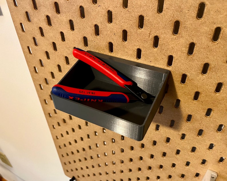 May include: A gray plastic tool holder with two red and blue pliers inside. The holder is mounted on a pegboard wall.