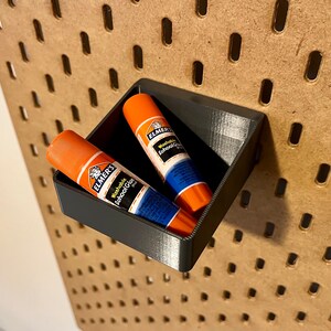 May include: Two orange and blue glue sticks are in a black plastic holder. The holder is mounted on a pegboard wall.