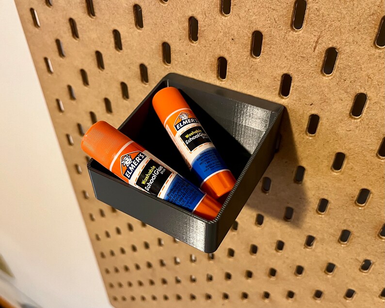 May include: Two orange and blue glue sticks are in a black plastic holder. The holder is mounted on a pegboard wall.