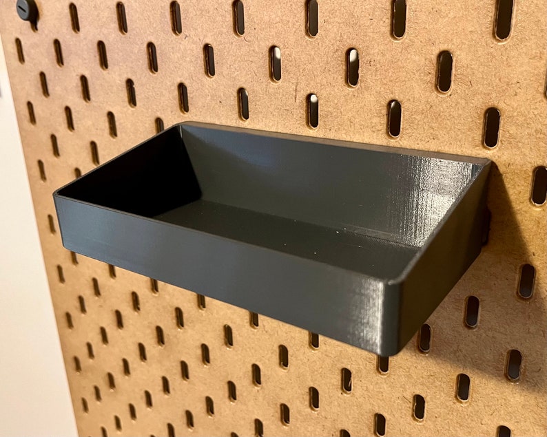 May include: A black plastic storage tray with a slightly angled front edge. The tray is mounted on a pegboard wall.