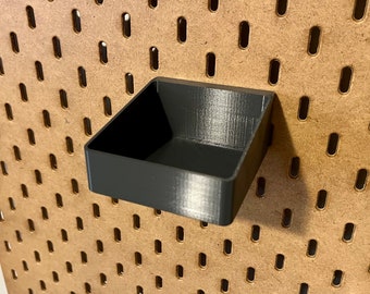 May include: A black plastic pegboard shelf with a square shape. The shelf is mounted on a brown pegboard.