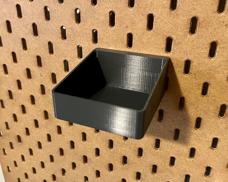 May include: A black plastic pegboard shelf with a square shape. The shelf is mounted on a brown pegboard.