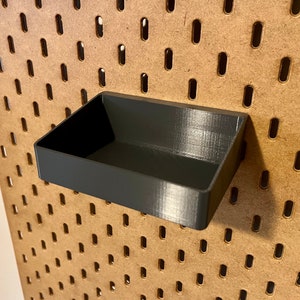 May include: A black plastic storage bin with a rectangular shape, designed to fit into a pegboard wall.