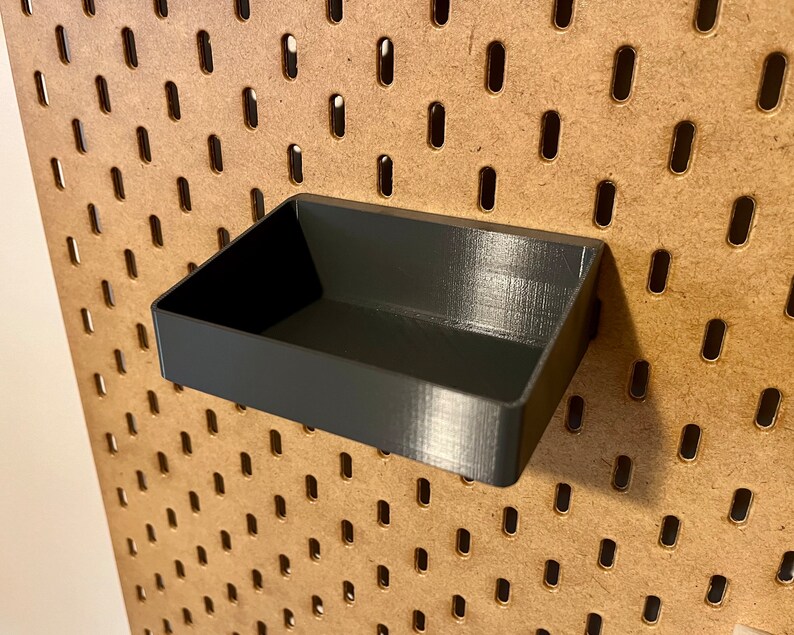 May include: A black plastic storage bin with a rectangular shape, designed to fit into a pegboard wall.