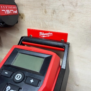 May include: A red and black Milwaukee digital tool with a screen and buttons, mounted on a wooden surface. The Milwaukee logo is visible on a red bracket. A black and red tape measure is partially visible.