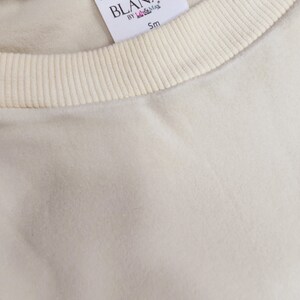 以下が含まれることがあります： クリーム色のリブ編みのネックラインが付いた白いコットン製のTシャツ。シャツには、ネックラインに縫い付けられた白いタグがあり、「Exclusively designed by BLANKXY BY lil & Max Sm」と書いてあります