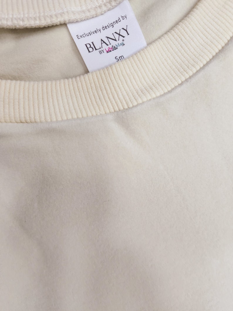 以下が含まれることがあります： クリーム色のリブ編みのネックラインが付いた白いコットン製のTシャツ。シャツには、ネックラインに縫い付けられた白いタグがあり、「Exclusively designed by BLANKXY BY lil & Max Sm」と書いてあります