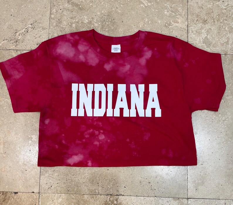 Pu&ograve; includere: Maglietta rossa con la scritta bianca "INDIANA". La maglietta ha un effetto tie-dye con macchie bianche. &Egrave; una maglietta a maniche corte e girocollo.