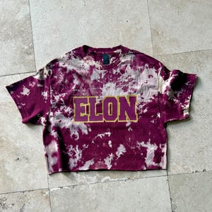 Pu&ograve; includere: Una t-shirt corta, tie-dye, nei toni del bordeaux e del bianco. La parola "ELON" &egrave; stampata sul davanti con un contorno dorato e un riempimento viola. La maglietta ha un girocollo e maniche corte.