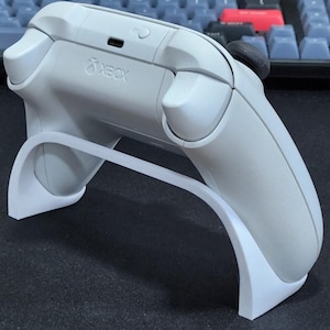 Puede incluir: Un mando Xbox blanco se muestra en un soporte blanco. El mando tiene un logotipo de Xbox negro y un botón negro en la parte superior.