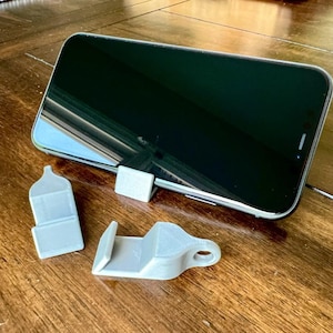 Op de afbeelding: Drie grijze plastic telefoonstandaards. Eén standaard is een klein vierkant, een ander is een hoog rechthoek en de derde is een gebogen vorm met een gat. Een zwarte smartphone staat op de vierkante standaard.