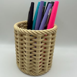 Puede incluir: Un porta lápices de mimbre tejido con un acabado marrón claro. El porta lápices está lleno de bolígrafos de varios colores, incluyendo rosa, morado, azul y verde.