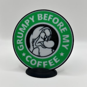 May include: A green and black round sign with a cartoon character of a grumpy dwarf. The sign says "Grumpy Before My Coffee" with a star on either side of the word "Coffee".