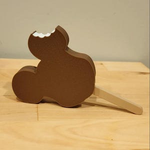 Può includere: Un gelato al cioccolato marrone a forma di Mickey Mouse su un bastoncino con un morso nella parte superiore.