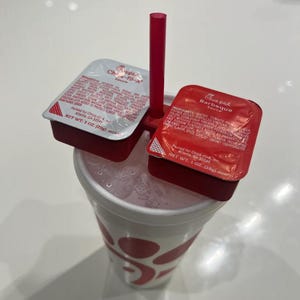 May include: A white Chick-fil-A cup with a red straw and two red condiment packets. The packets are labeled "Chick-fil-A Sauce" and "Barbeque Sauce".