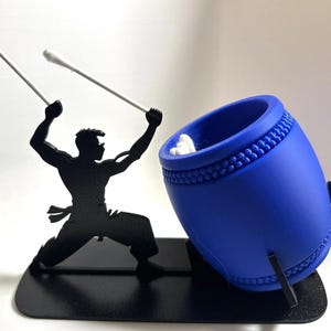 May include: A black silhouette of a person holding two white cotton swabs, striking a blue drum with a white cotton swab inside. The drum is on a black stand.