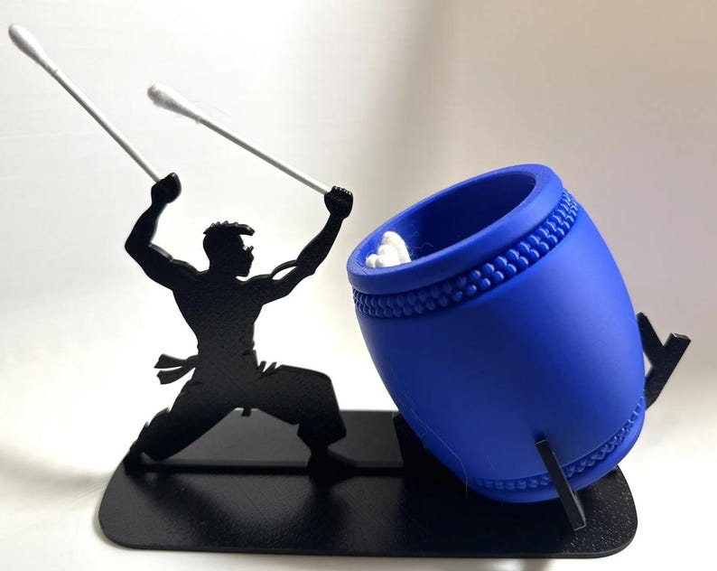 May include: A black silhouette of a person holding two white cotton swabs, striking a blue drum with a white cotton swab inside. The drum is on a black stand.