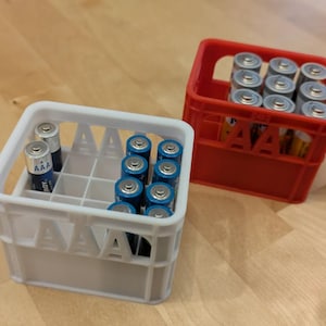 Puede incluir: Dos cajas de plástico para guardar pilas AAA. Una caja es blanca y la otra es roja. Ambas cajas tienen las letras "AAA" impresas en la parte delantera.