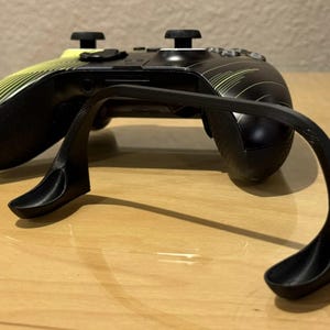 May include: A black and yellow Xbox controller with a black plastic stand. The stand is designed to hold the controller upright.