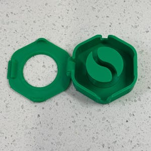 May include: A green plastic container with a hinged lid. The container has a round opening and a small green plastic insert with a curved shape inside.