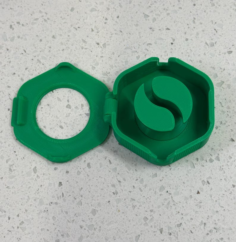 May include: A green plastic container with a hinged lid. The container has a round opening and a small green plastic insert with a curved shape inside.