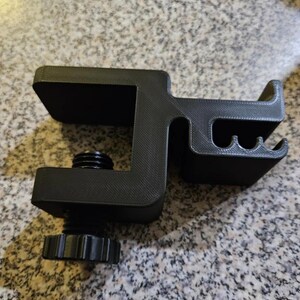 Puede incluir: Una abrazadera negra, impresa en 3D, con un mecanismo de tornillo y una superficie texturizada. La abrazadera tiene forma de C con una parte superior plana y una secci&oacute;n curva con tres ranuras peque&ntilde;as. El tornillo tiene una perilla moleteada para un ajuste f&aacute;cil.