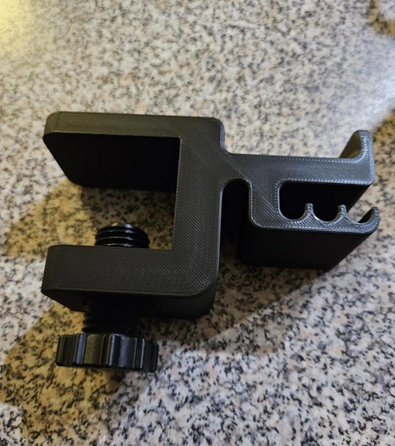 Puede incluir: Una abrazadera negra, impresa en 3D, con un mecanismo de tornillo y una superficie texturizada. La abrazadera tiene forma de C con una parte superior plana y una secci&oacute;n curva con tres ranuras peque&ntilde;as. El tornillo tiene una perilla moleteada para un ajuste f&aacute;cil.