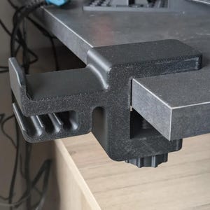 Puede incluir: Una abrazadera de escritorio negra, dise&ntilde;ada para sujetarse a un escritorio o mesa. La abrazadera tiene una superficie texturizada y un mecanismo de tornillo para apretar. El dise&ntilde;o incluye una caracter&iacute;stica en forma de gancho para sujetar objetos.