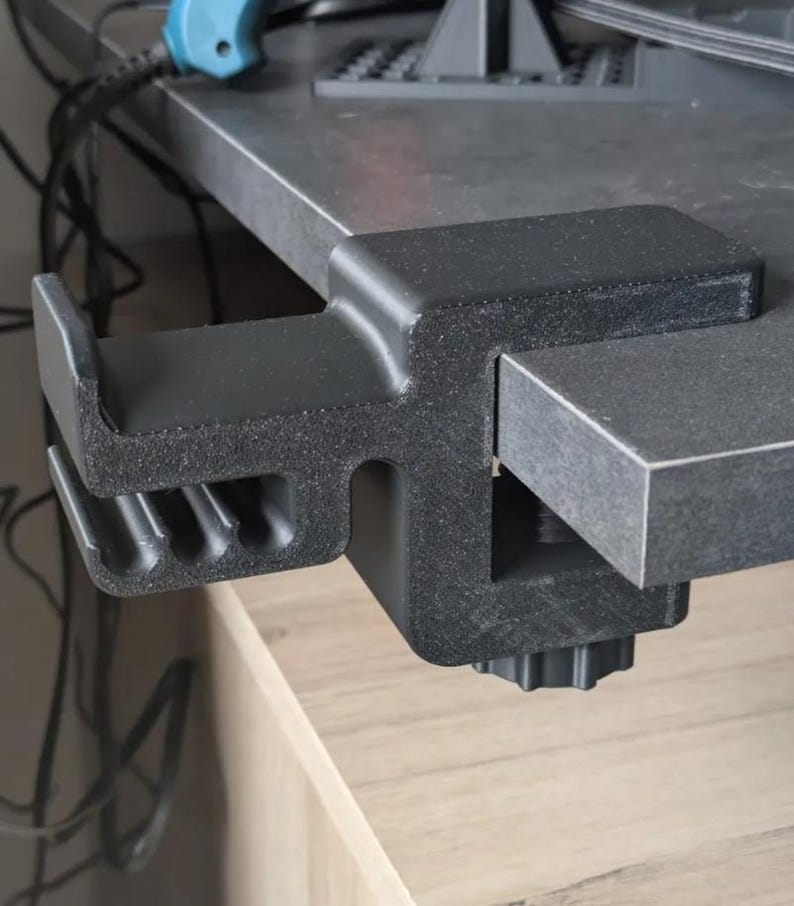 Puede incluir: Una abrazadera de escritorio negra, dise&ntilde;ada para sujetarse a un escritorio o mesa. La abrazadera tiene una superficie texturizada y un mecanismo de tornillo para apretar. El dise&ntilde;o incluye una caracter&iacute;stica en forma de gancho para sujetar objetos.
