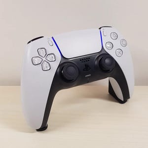 Op de afbeelding: Een witte en zwarte PlayStation 5 DualSense draadloze controller met een blauwe lichtbalk. De controller heeft een zwart touchpad, twee zwarte analoge sticks en een zwart D-pad.