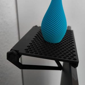 May include: A black metal shelf with a honeycomb design, mounted on a black surface. A blue, spiraled vase sits on the shelf.