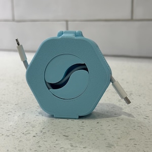 May include: A light blue cable organizer with a hexagonal shape and a circular opening in the center. The cable is white and is partially wound around the organizer.