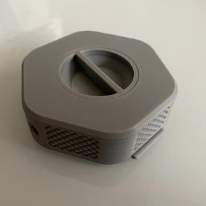 May include: A gray hexagonal-shaped filter holder with a small, rectangular opening in the center. The filter holder has a textured pattern on the sides.
