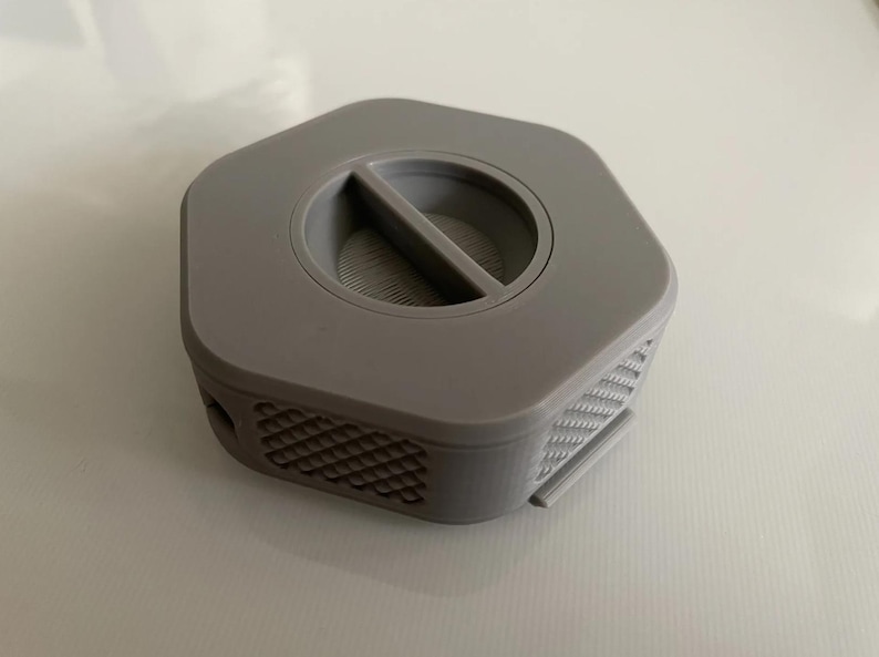 May include: A gray hexagonal-shaped filter holder with a small, rectangular opening in the center. The filter holder has a textured pattern on the sides.