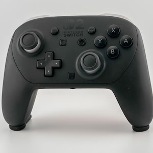 Pode incluir: Um comando Nintendo Switch Pro preto com detalhes brancos. O comando possui uma cruz direcional, manípulos analógicos e botões marcados X, Y, A e B. O logótipo Nintendo Switch é visível.
