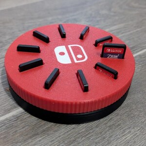 May include: A red and black Nintendo Switch game cartridge holder. The circular holder has slots for game cartridges and features the Nintendo Switch logo. One cartridge is labeled "Zelda".
