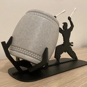 May include: A gray ceramic drum with a black metal stand and a silhouette of a person holding drumsticks. The stand has a base and two arms that hold the drum.
