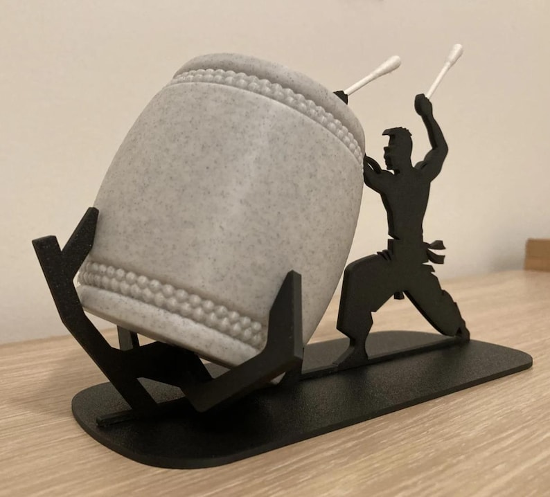 May include: A gray ceramic drum with a black metal stand and a silhouette of a person holding drumsticks. The stand has a base and two arms that hold the drum.
