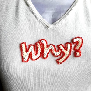 May include: White v-neck shirt with the word "Why?" spelled out in red and white sequins.