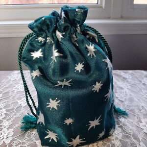 May include: A teal drawstring pouch featuring a repeating pattern of silver stars. The bag is made of a shimmering fabric and includes dark green twisted cord drawstrings with small tassels at the ends to secure the top opening of the pouch.