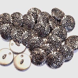 Peut inclure: Une collection de boutons ronds en métal présentant un motif floral et de volutes en relief de couleur argentée sur le devant. Le revers des boutons est lisse et métallique, chacun étant muni d'une petite boucle centrale pour la fixation.