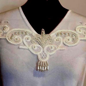 May include: A white sleeveless top with a V-neck, embellished with a cream-coloured embroidered design. The embroidery includes swirling patterns, beaded details, and a dangling fringe, creating an elegant decorative element.