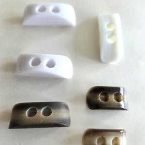 May include: A collection of six rectangular buttons with two holes each. Two buttons are solid white with a curved profile, one is a translucent white, and three are a gradient of dark brown to light beige with a smooth, glossy finish.